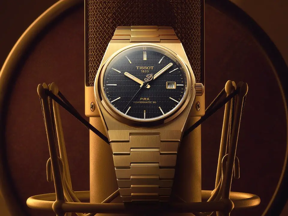 Eine Tissot PRX Automatik-Uhr in Gold ist auf einem Mikrofon platziert, vor einem sanften, goldenen Hintergrund, der eine elegante Atmosphäre schafft.