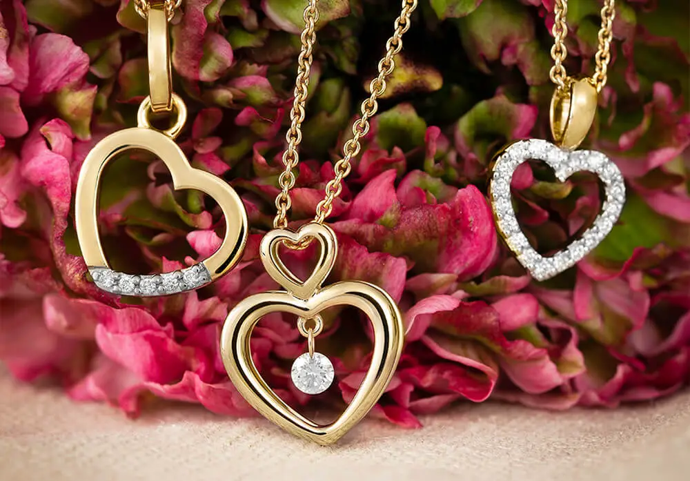 Drei goldene Herzanhänger mit zarten Ketten, ein Anhänger hat glitzernde Steine, vor einem Hintergrund aus rosa Blüten.
