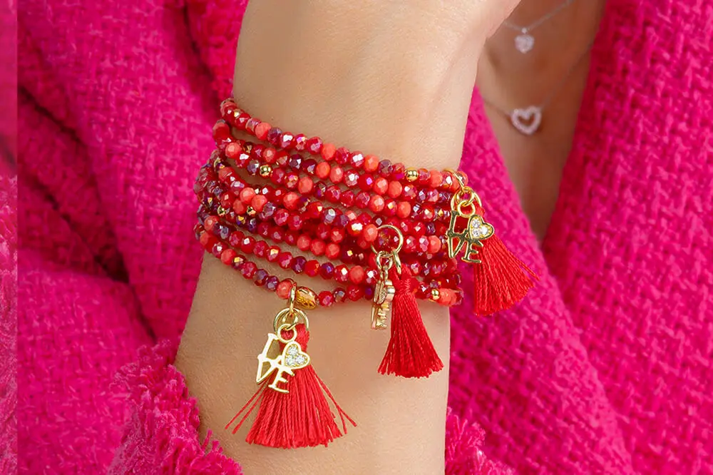 Eine Hand trägt mehrere rote Armbänder aus Perlen mit goldenen Anhängern und roten Quasten, vor einem pinken Hintergrund.