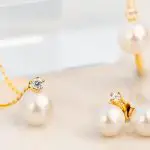 Ein goldener Halsreifen und Ohrringe mit kleinen Perlen und funkelnden Zirkonia-Steinen auf einem hellen, glatten Hintergrund. 