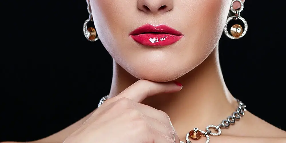 Eine Frau mit elegantem Make-up und rotem Lippenstift posiert mit einer Hand an ihrem Hals, der eine auffällige Kette mit Schmuck trägt.